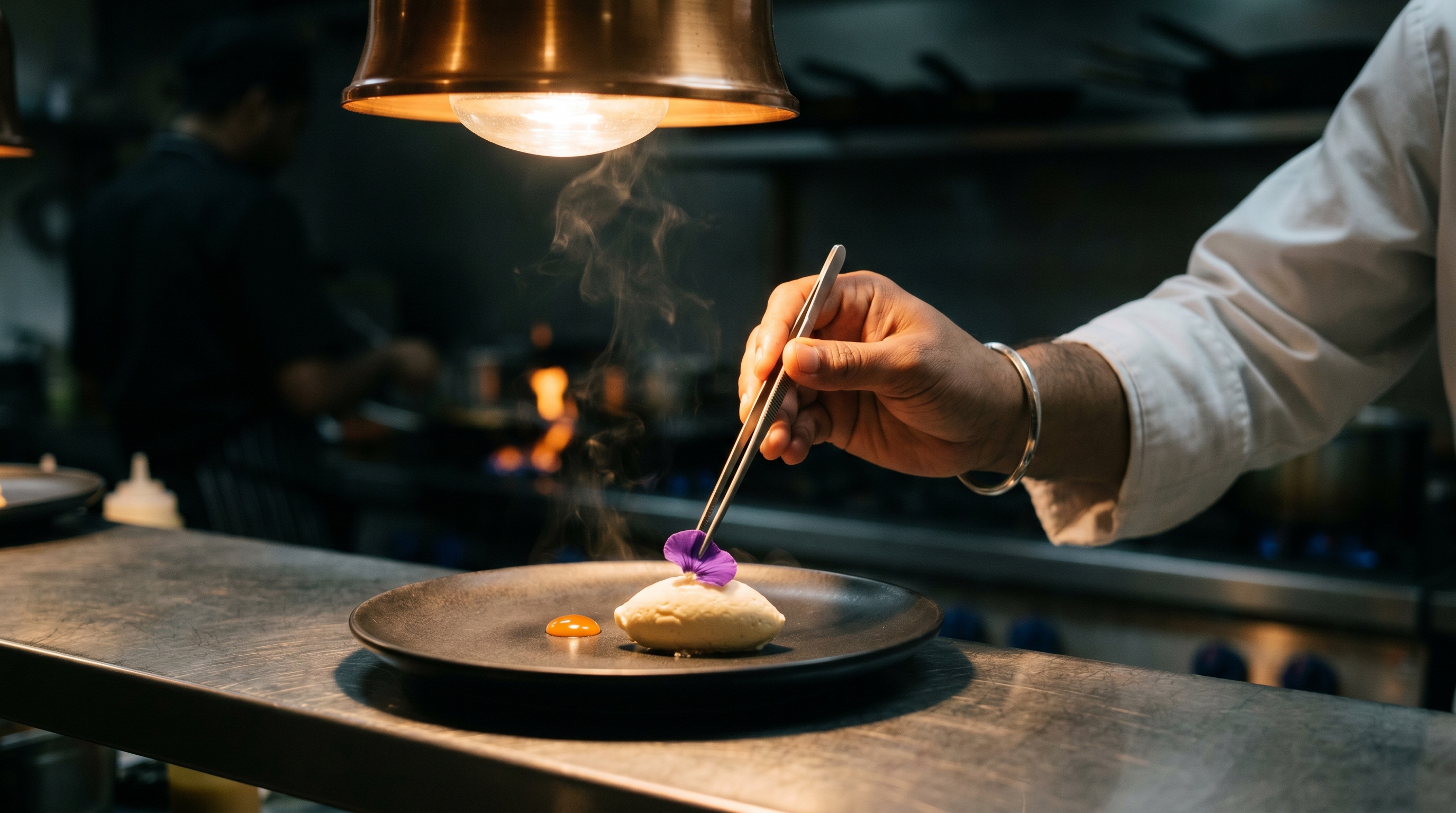 An overhead view of a beautifully composed Indian fine-dining tasting-menu pass with a chef's hand finishing a plate