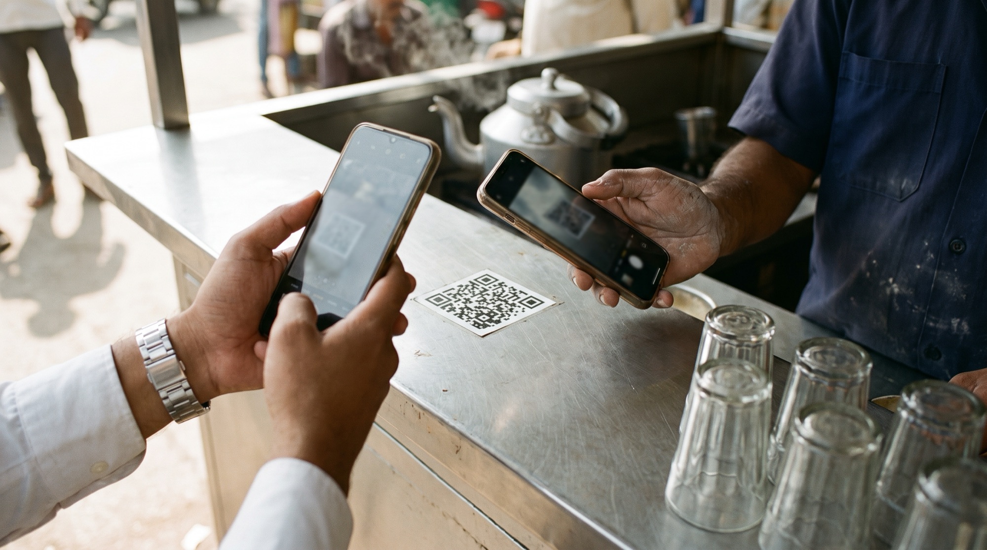 Two hands at a Mumbai chai stall counter, one in a white shirt cuff with a steel watch and one in a navy work shirt, both holding phones over the same QR code sticker
